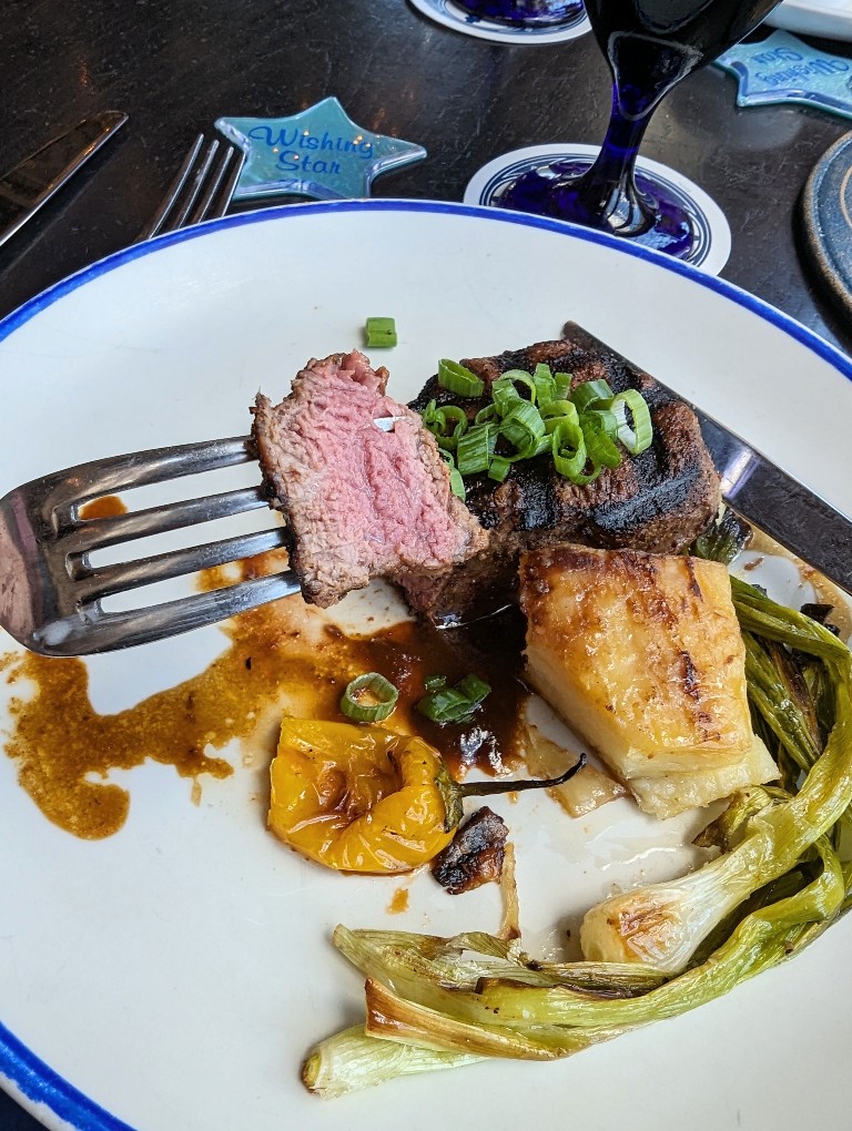 A bite of medium steak on a fork with a plate of potato and vegetables behind