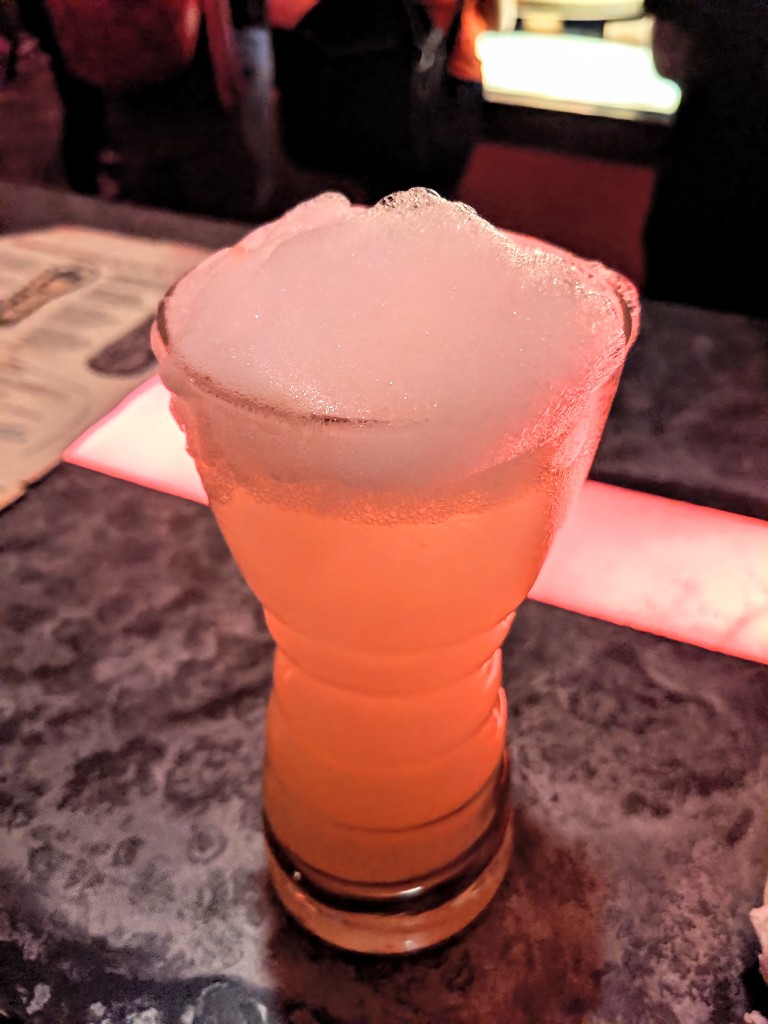 A peachy-orange colored drink with tall white foam sits on the Oga's Cantina bar