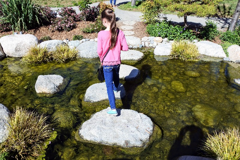 Girl crosses water on large rocks when visiting Balboa Park with kids.