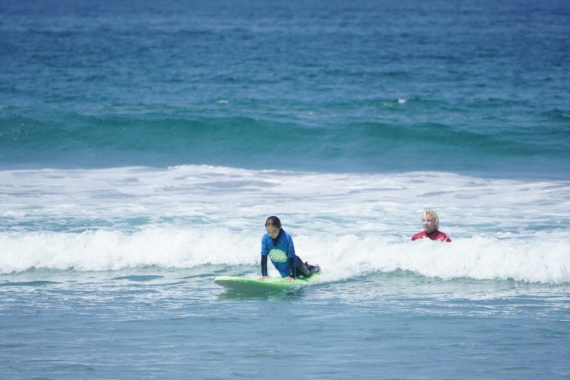 Girl prepares to stand on surf board while Pacific Surf School instructor looks on