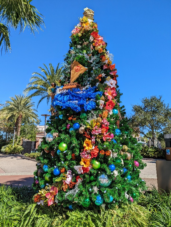 Tropical flowers ring a large Christmas tree with Moana's boat as a focal point at Disney Springs Christmas Tree Stroll
