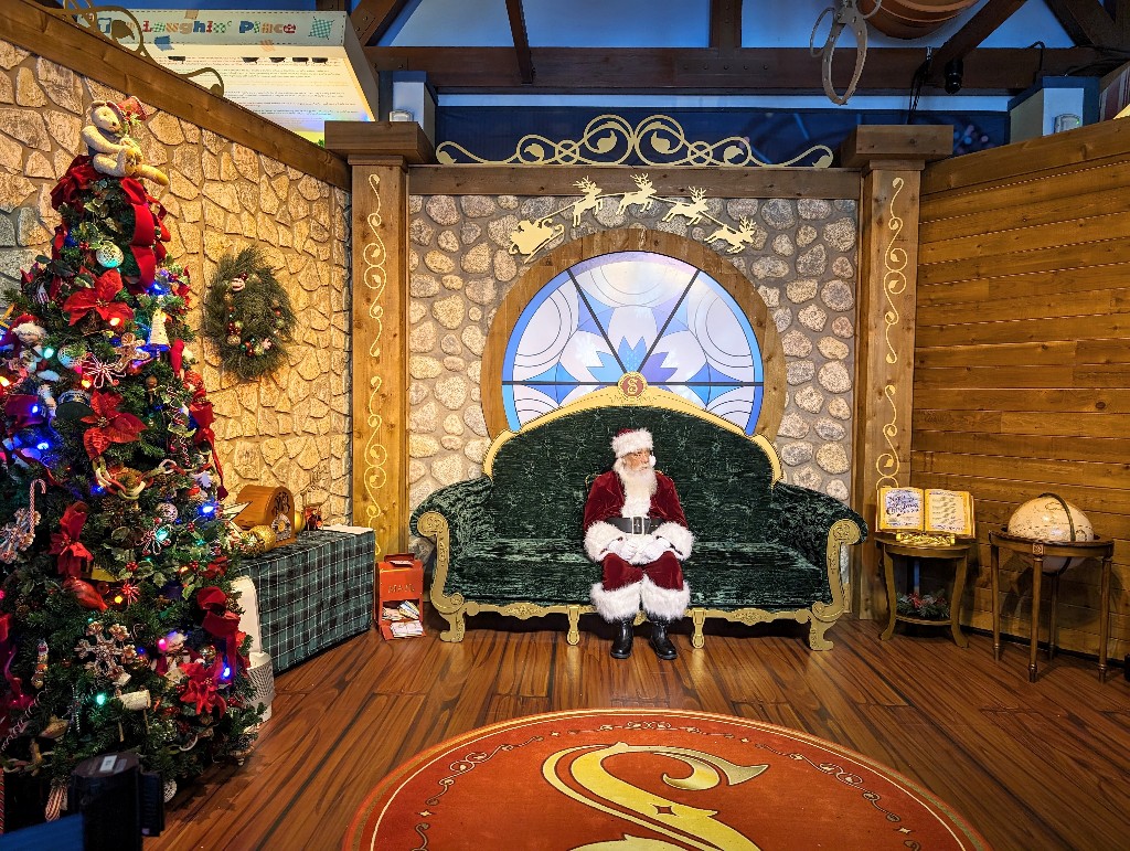 Santa sits on a large green velvet couch with lovely Christmas decorations all around waiting for his next visitor at Once Upon a Toy in Disney Springs