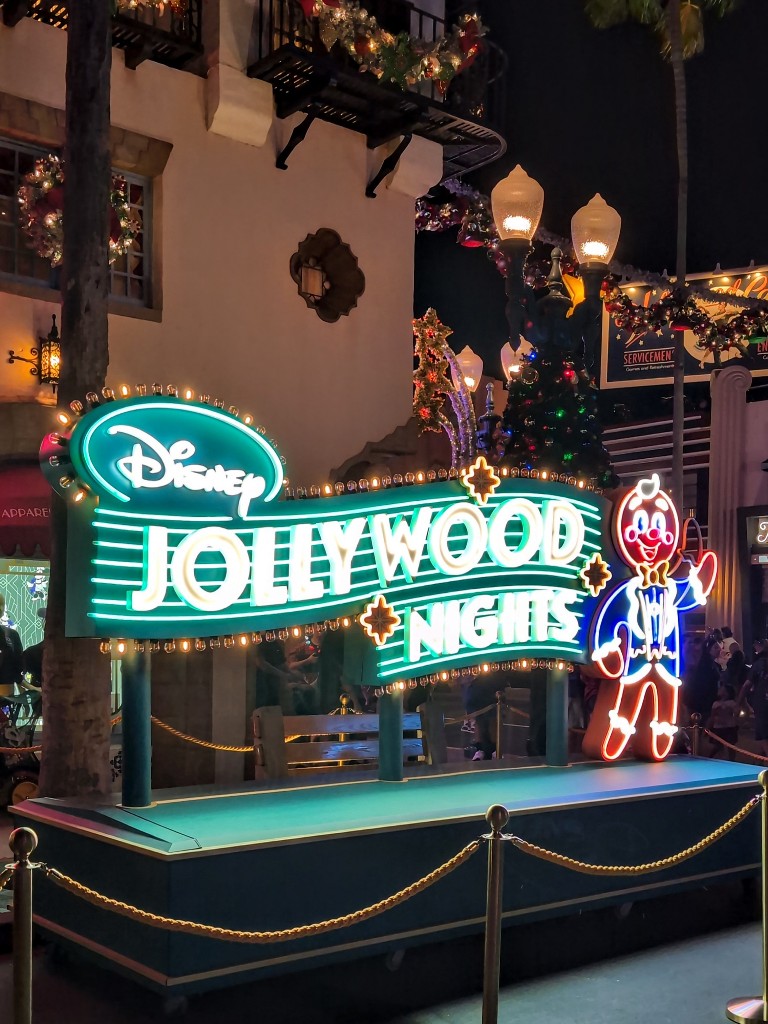 A teal neon sign says "Disney Jollywood Nights" with a neon gingerbread man next to it at Hollywood Studios during Christmas