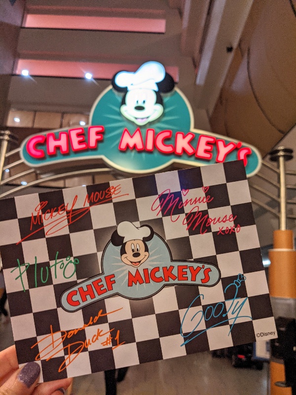 Chef Mickey's sign with woman holding Chef Mickey autograph card souvenir provided as part of the meal