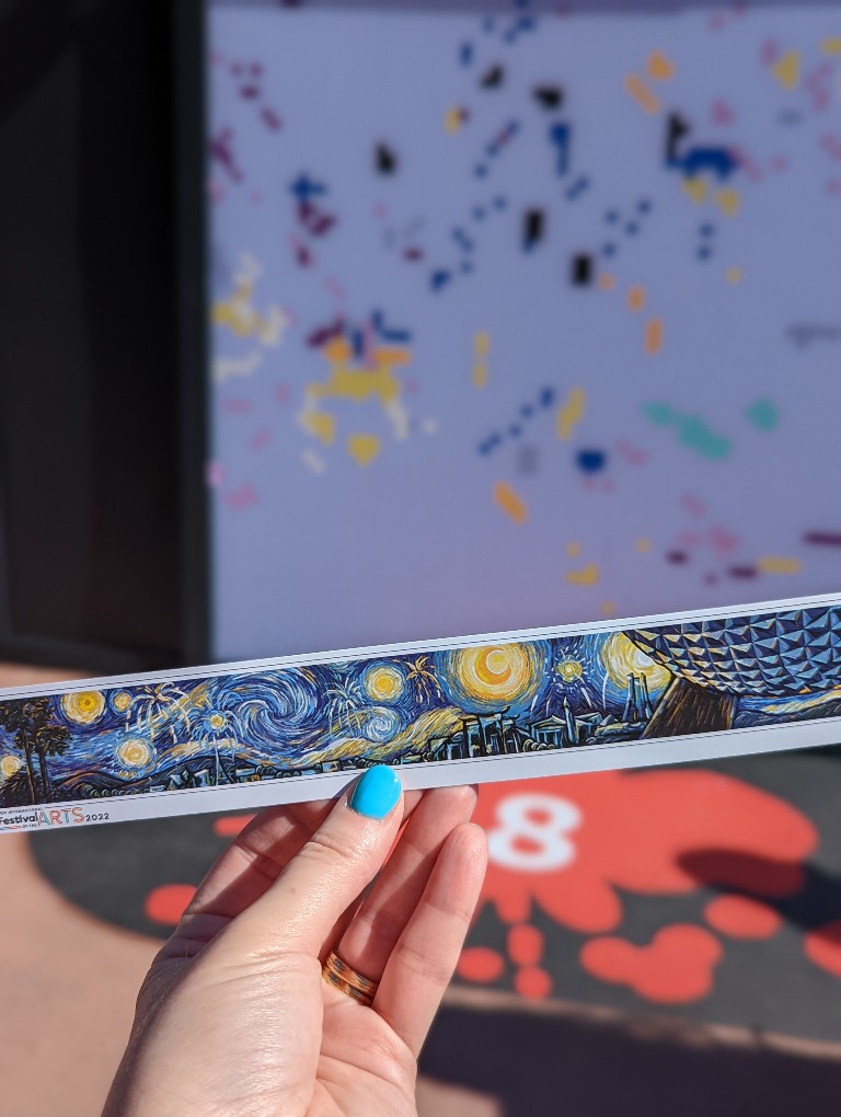 Woman holds a paper bookmark of the finished Expression Section: A Paint-by-Number Mural at Epcot Festival of the Arts