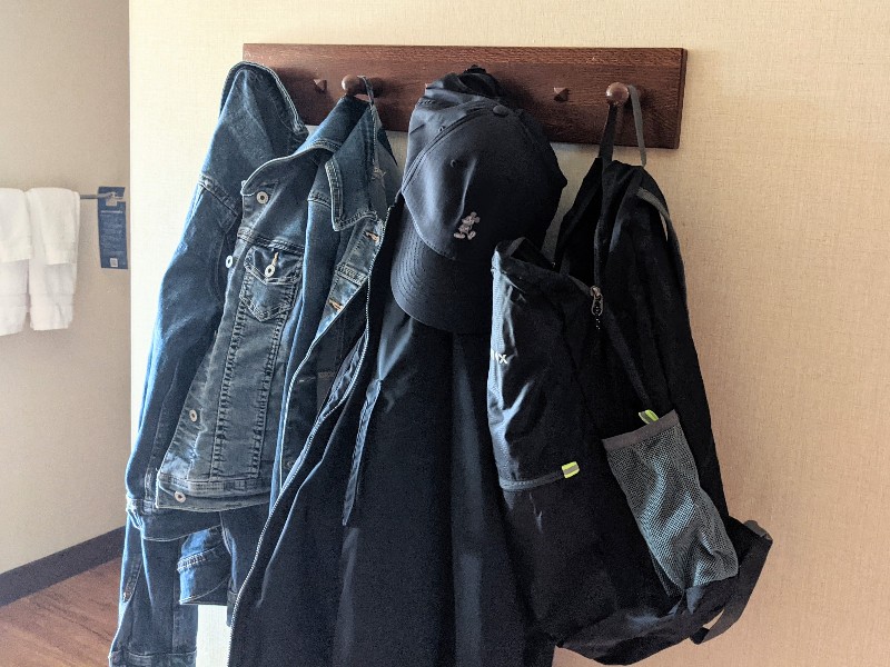 A black packable backpack hangs on a hook next to jackets and a Mickey Mouse hat in a Disney hotel room.