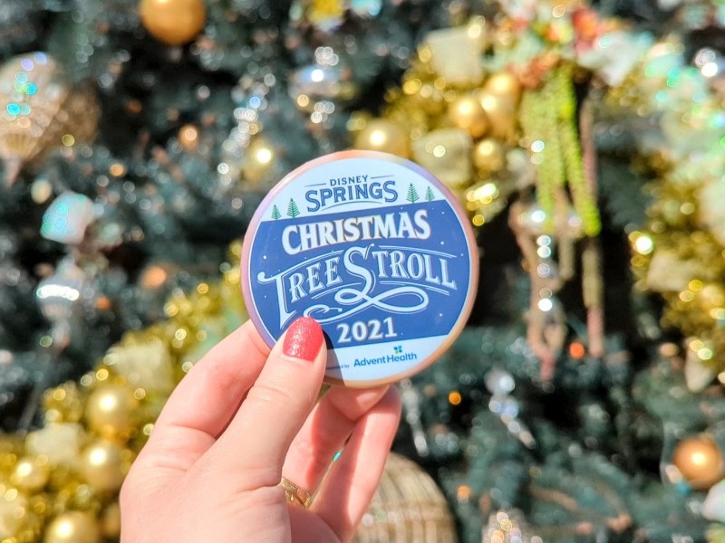 Woman holds Disney Springs Christmas Tree Stroll pin in front of a sparkling Christmas tree