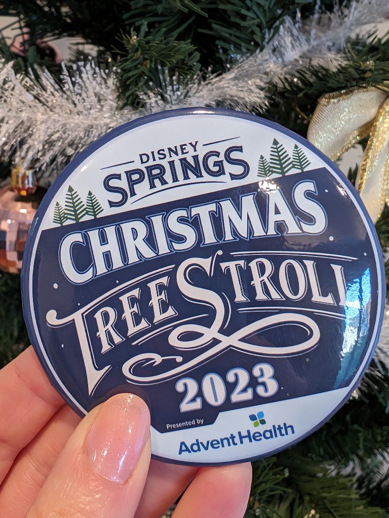 Disney Springs Christmas Tree Stroll 2023 prize large metal pin held in front of a Christmas tree