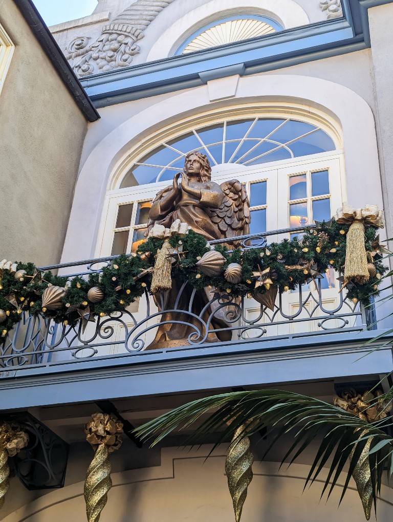 An angel serenely stands on a balcony adorned with greenery and gold embellishments in New Orleans Square