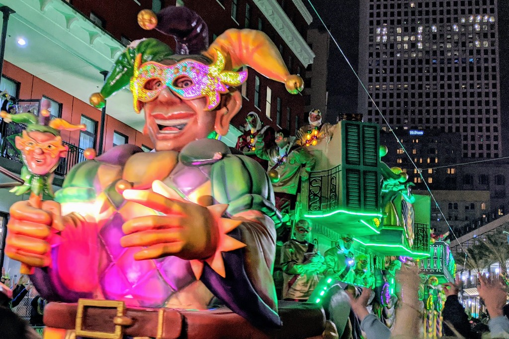 A large joker float illuminated along a nighttime Mardi Gras parade route