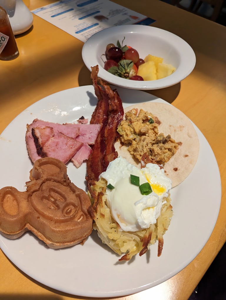 A buffet plate with steak and eggs on a tortilla, bacon, smoked ham, Mickey waffle and fresh fruit
