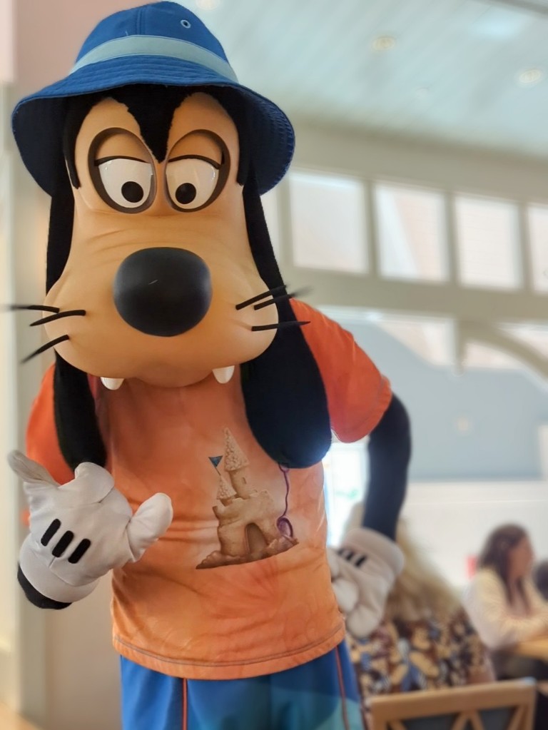 Goofy makes a hang loose sign in his bucket hat, rash guard, and swim trunks at Cape May Cafe