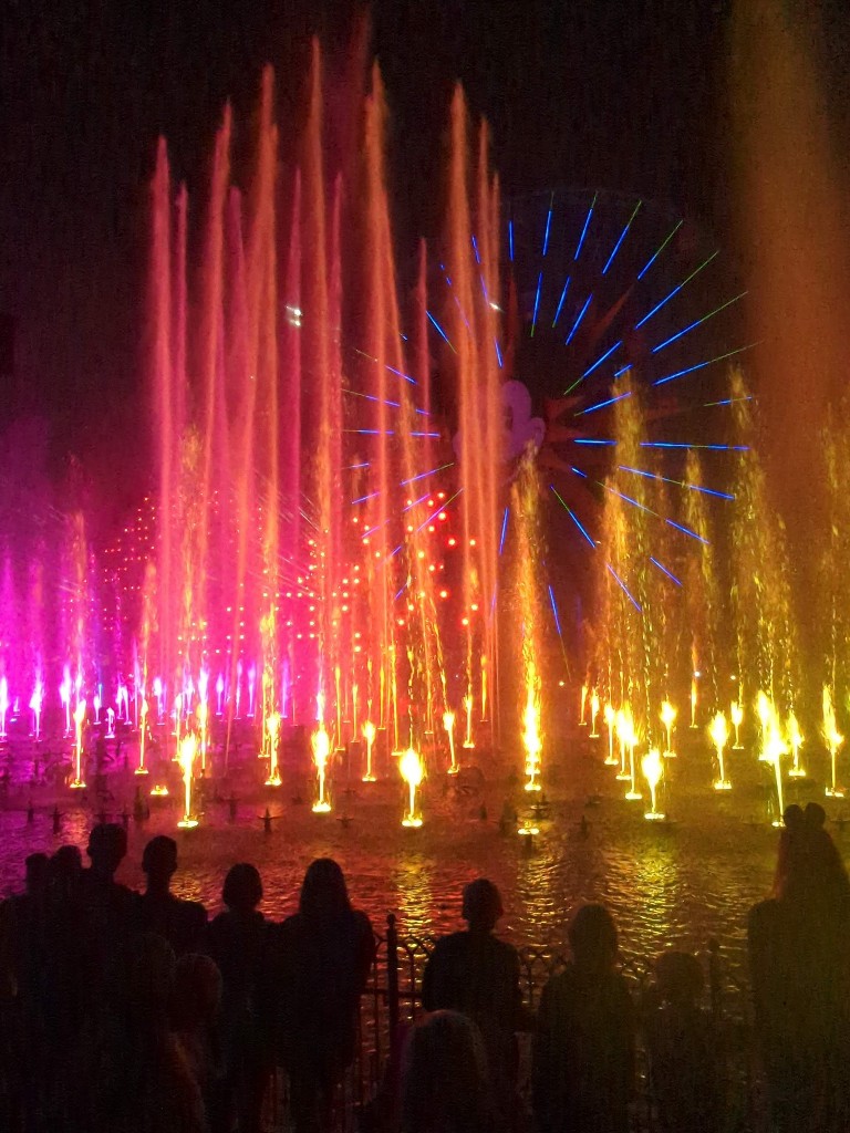 Pink, orange and yellow lights illuminate spouts of water in the World of Color virtual queue viewing area