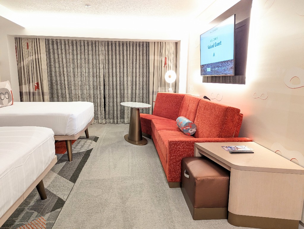Subtle mask wallpaper accent wall with a bright orange couch that folds down to a single bed and two queen beds provide comfortable sleeping for 5 guests at Disney's Contemporary Resort Garden Wing