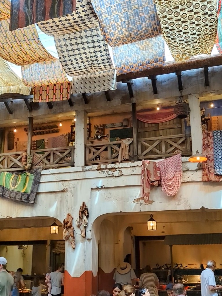 The Tusker House buffet line is surrounded by a distressed two story building with gorgeous wooden railings, tapestries, and overhead cloths that act as a canopy mimicking a African marketplace