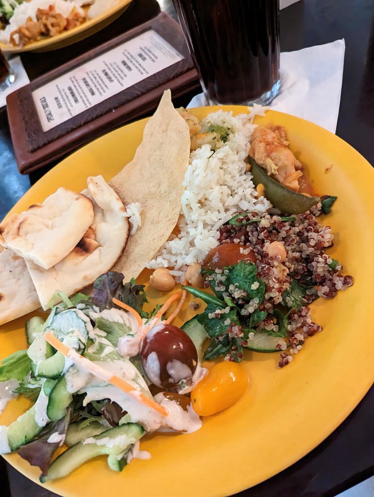 A bright yellow plate heaped with salad, cous cous salad, rice, curries, and naan