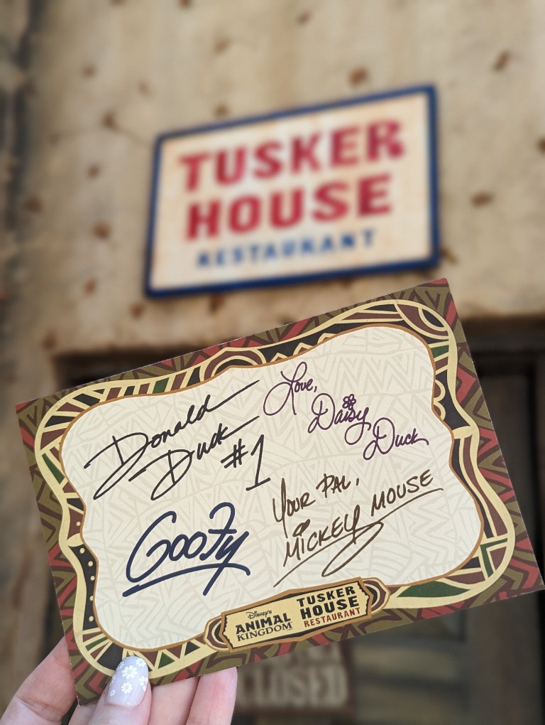 A signed card with character autographs is held in front of a Tusker House sign