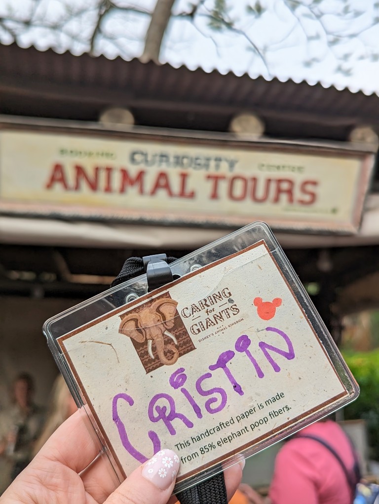 A Caring for Giants tour name tag with the name "Cristin" is held up in front of the Curiosity Animal Tour kiosk