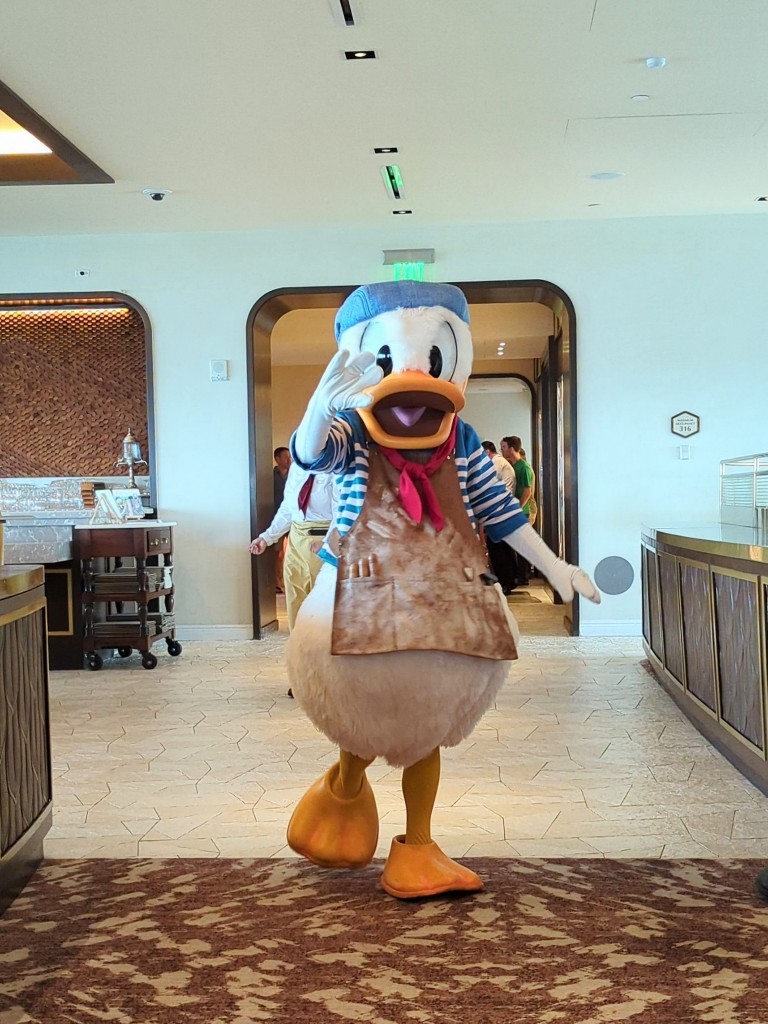 Donald Duck waves to guests as he enters Topolino's Terrace wearing a clay dusted smock and blue striped shirt