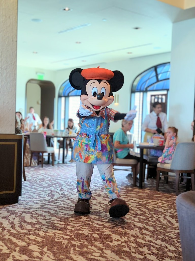 Mickey welcomes guests with open arms dressed in a striped shirt and paint spattered smock at Topolino's Terrace character breakfast