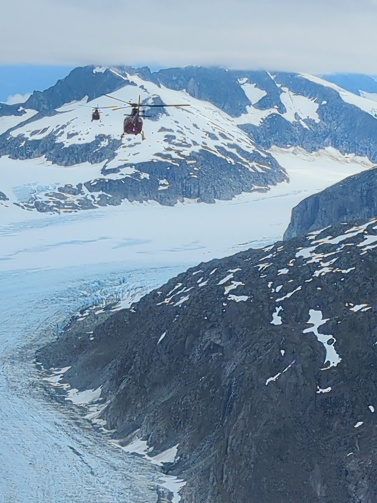 Two helicopters fly over Mendenhall Glacier with snowcapped mountains on each side