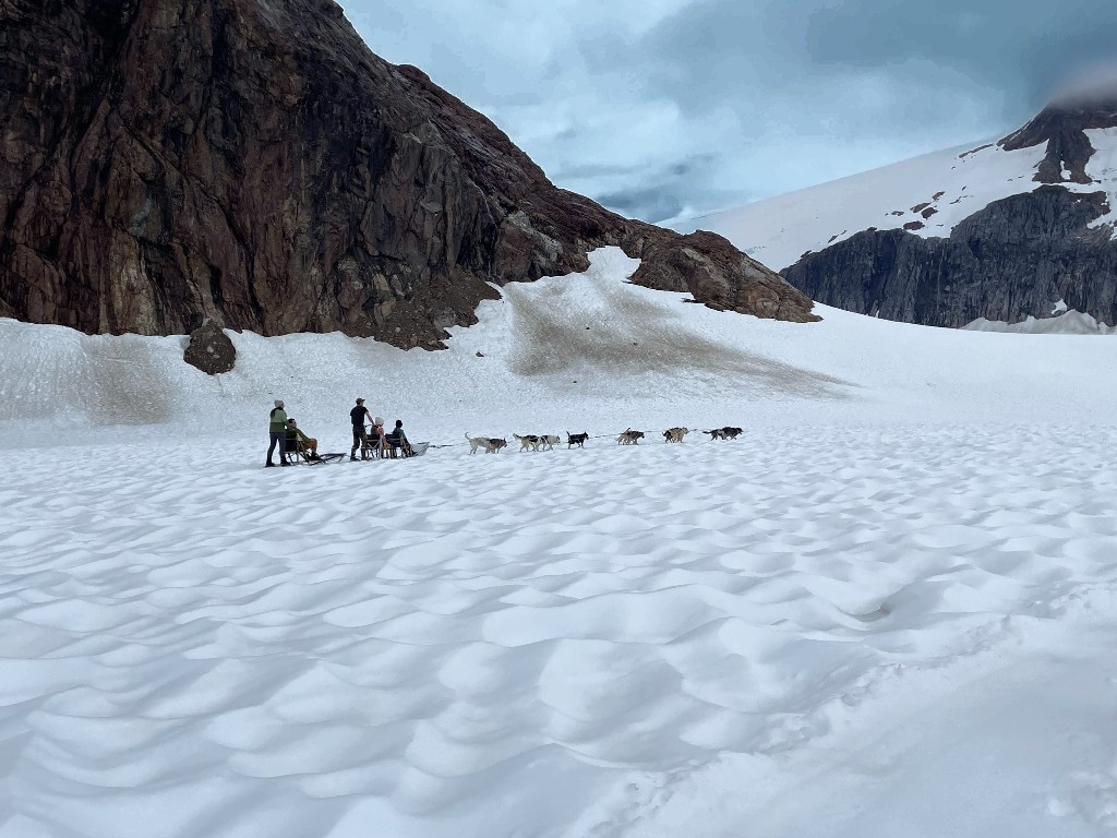 Rocky cliffs in the background and a sled dog team with guests on a endless swath of snow during the Dog Sledding on Mendenhall Glacier Disney Alaska Cruise excursion