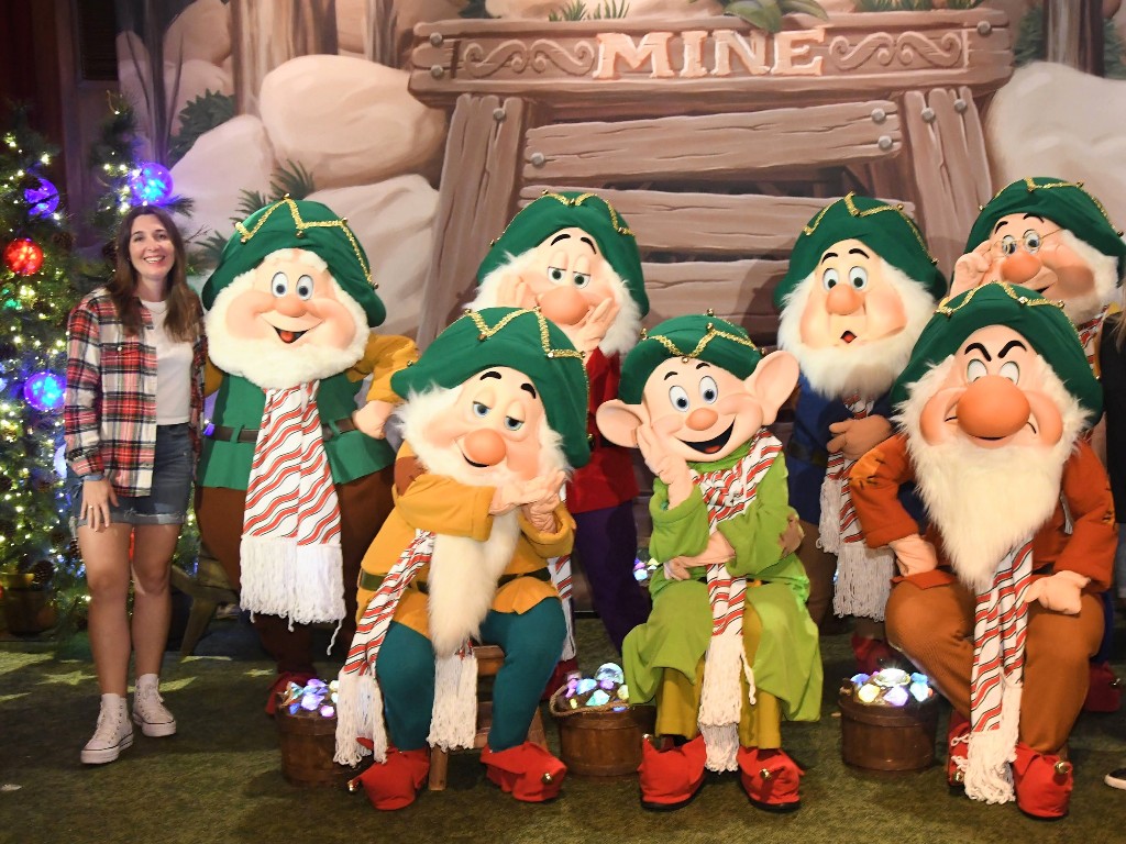 Woman in a festive plaid shirt and shorts stands next to the seven dwarfs who are all wearing striped scarves and jolly green hats during Mickey's Very Merry Christmas Party