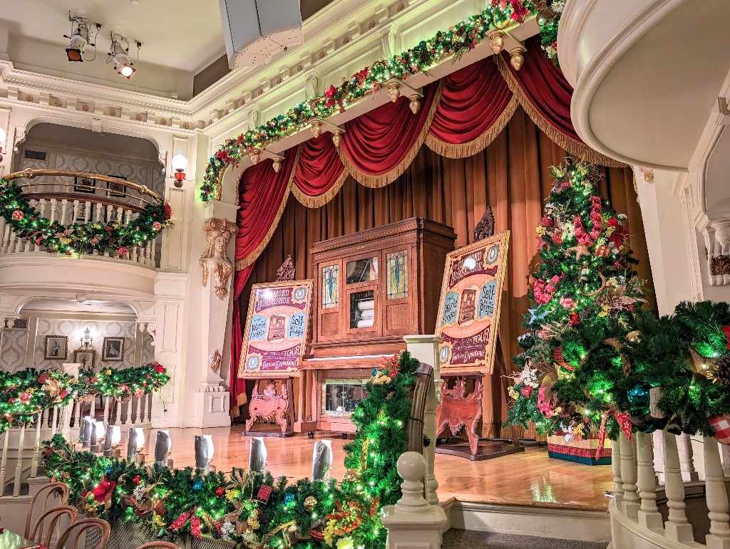 Diamond Horseshoe Christmas decorations adorn the railings and a large tree with red accents is a focal point of the player piano stage