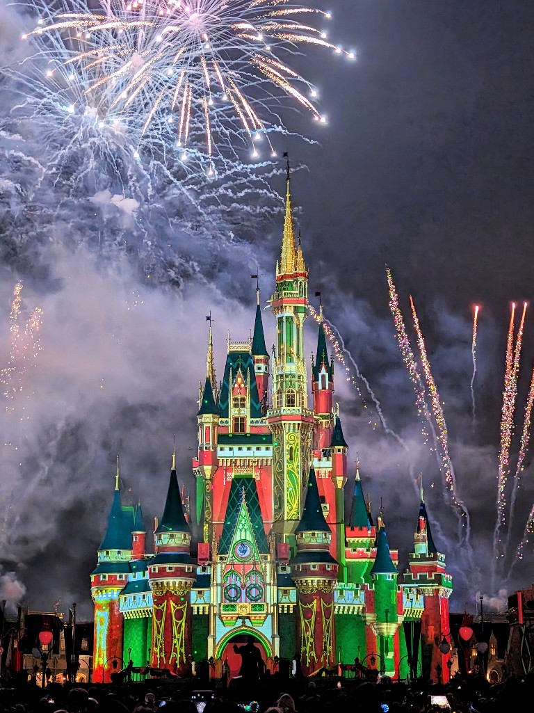 Cinderella Castle with green, red, and gold projections and fireworks in the background at MVMCP