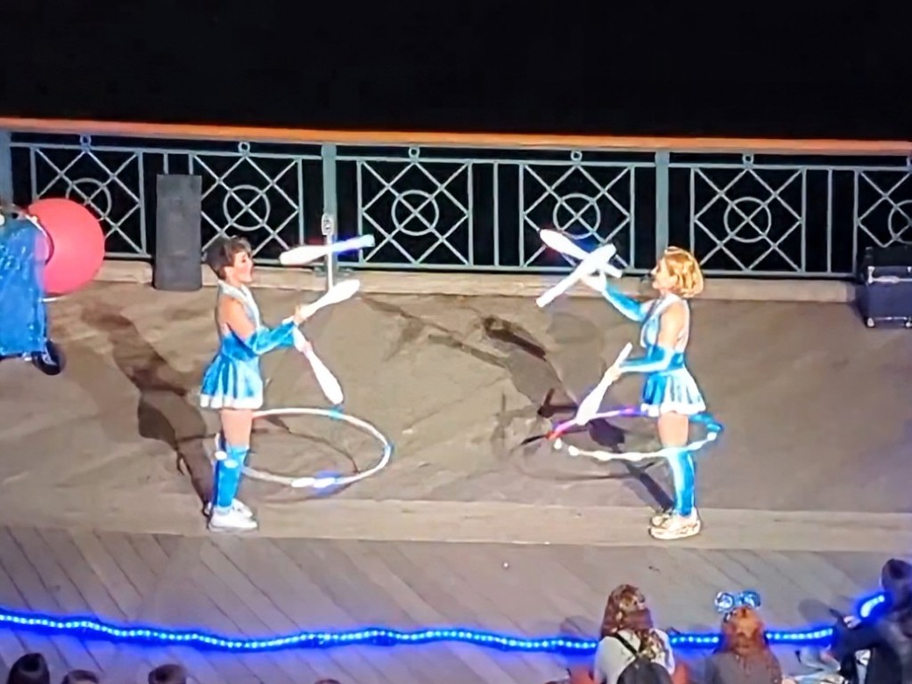 Two women entertain guests at Disney's Boardwalk in sparkling blue outfits by hula hooping while juggling 