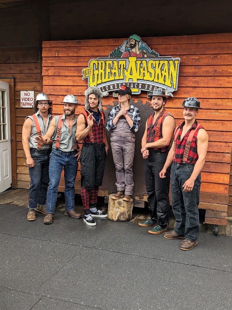 A group of lumberjacks and the host pose for photos under a large Great Alaskan Lumberjack Show sign