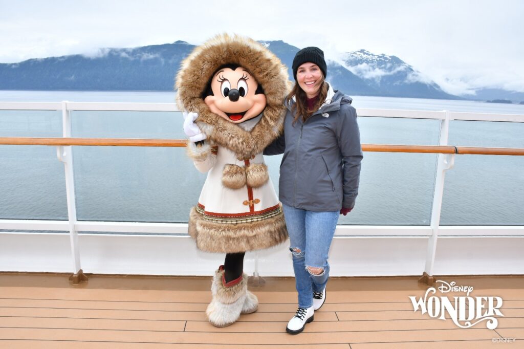 A woman wearing a winter hat, TriClimate coat, gloves, and waterproof boots poses with Minnie Mouse in front of a cloud covered Alaskan shore