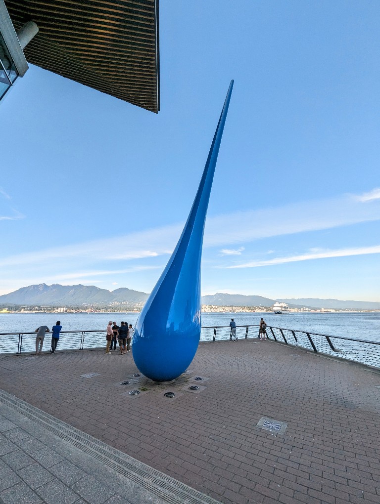 A large, bright blue water drop sculpture is framed by the North Mountains in Vancouver