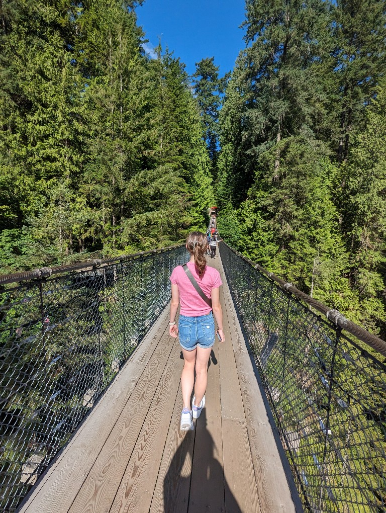 A girl in shorts and a t-shirt walks across Capilano Suspension Bridge with gorgeous pines in the background