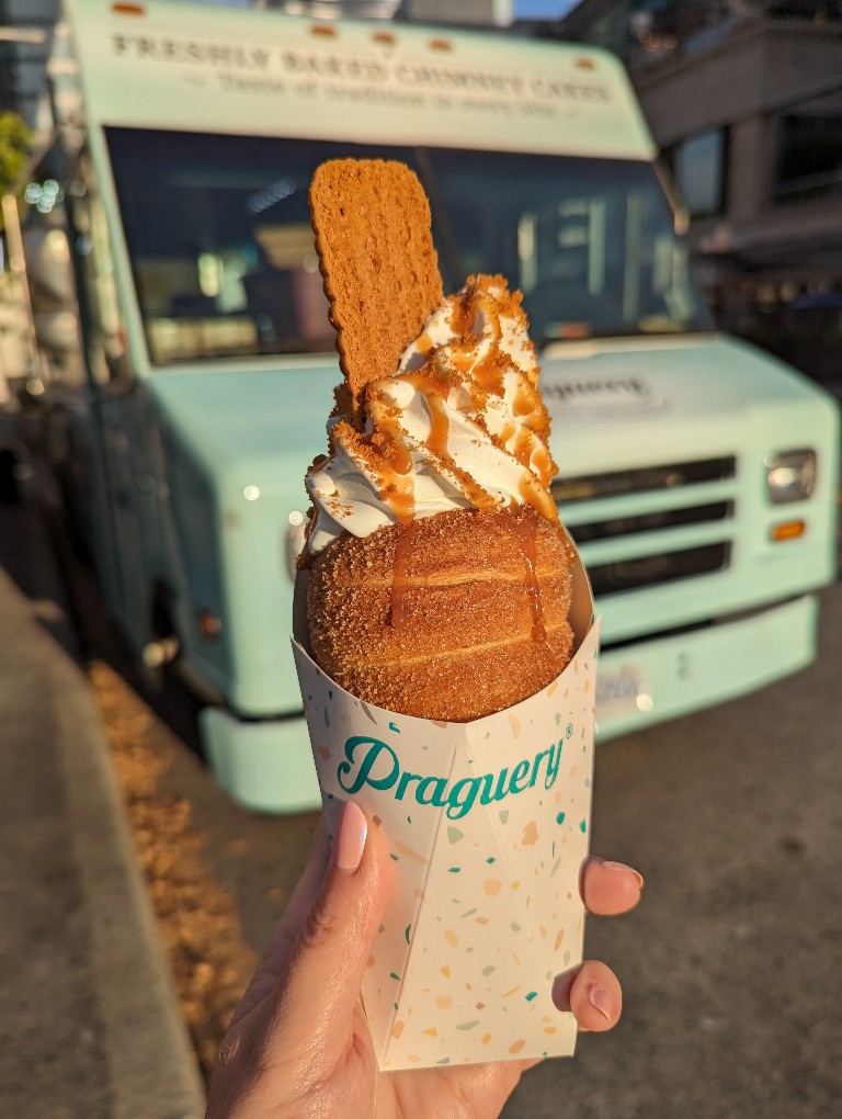 A delicious chimney cake filled with swirls of vanilla ice cream and drizzled with caramel is held in front of a blurry food truck