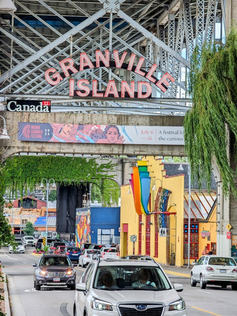 A large "Granville Island" sign hangs above and colorful murals adorn the buildings along the sides of the main street leading into this popular tourist destination