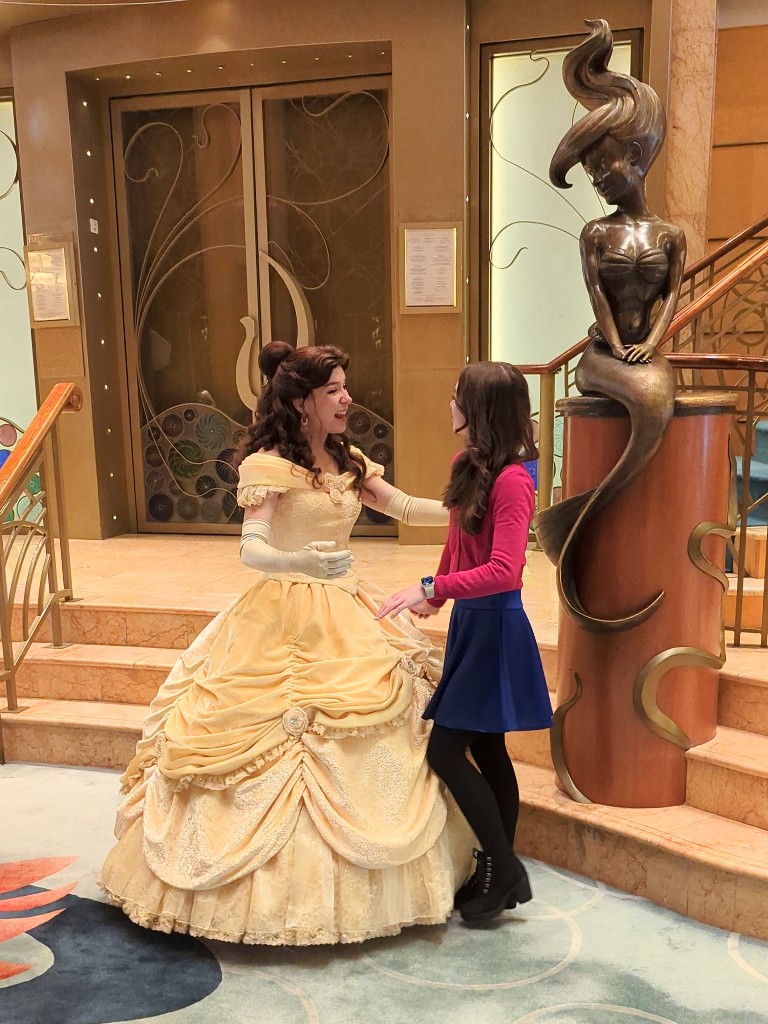 A teenage girl Disneybounds as Anna in a magenta cardigan, blue skirts, and black boots while meeting Belle on Disney Wonder
