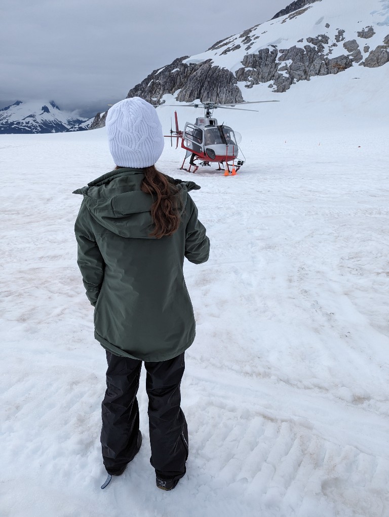 A teenage girl stand on Mendenhall Glacier snow pack in waterproof boots, pants, NorthFace jacket, and warm beanie with a helicopter in the background