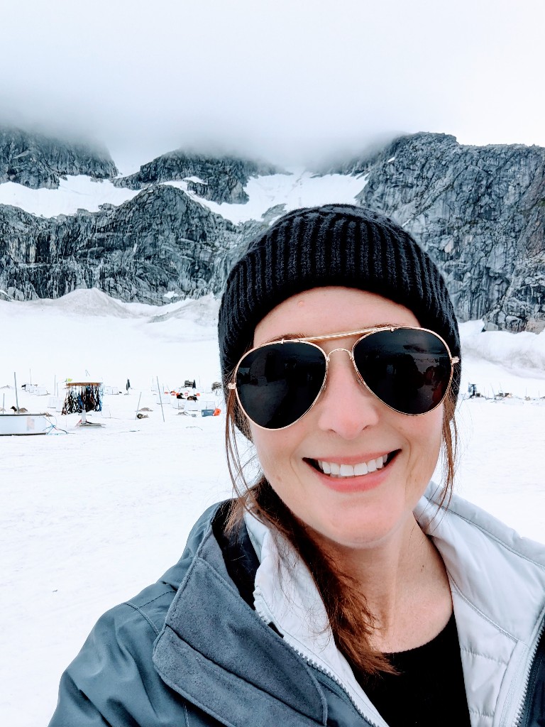 A woman smiles wearing sunglasses and a warm beanie at the Mendenhall Glacier dog sled camp