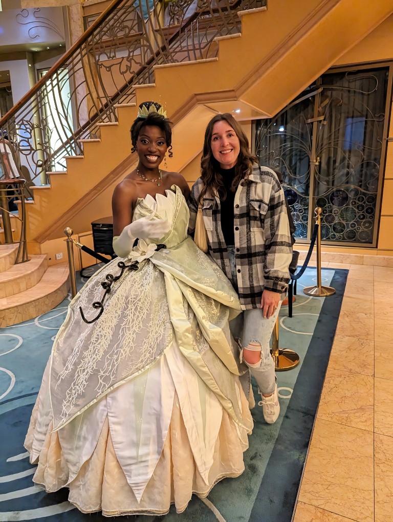 A woman poses with Tiana in jeans, tennis shoes, a black mockneck shirt and oversized plaid on a Disney Alaska Cruise